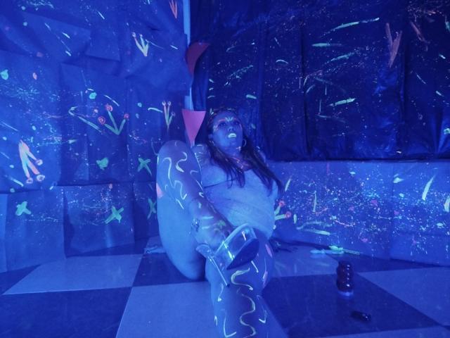 LexyDom is kneeling on a checkered floor, glowing neon paint on her body, tits exposed, in a blue-lit room, with a dildo on the floor to her right.