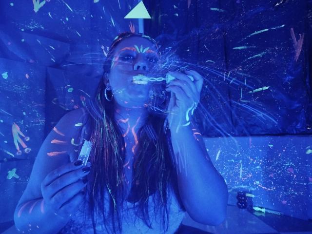 LexyDom, avec des motifs néon bleus et verts lumineux sur son visage et sa poitrine, tenant une cigarette éclairée au néon, portant des boucles d'oreilles pendantes, avec de longs cheveux bruns mouillés, dans une pièce éclairée en bleu foncé..