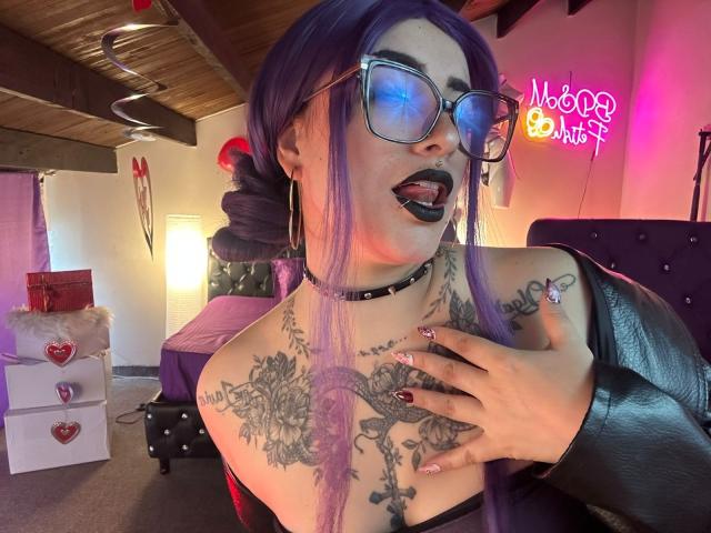 LilithDark69 with purple hair, black choker, tattoos, glasses, tongue out, black lipstick, hand on chest, leather sleeve, neon lights, and a sexy, goth, punk-inspired look.