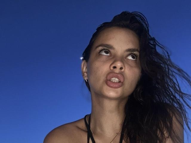 CourtneyBrunkhardt has wet, dark brown hair, puffy lips, and a surprised expression, looking up with brown eyes, wearing a black halter top, against a blue background.