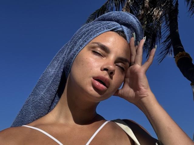 Courtney Brunkhardt con una toalla azul en la cabeza, ojos cerrados, boca ligeramente abierta, mano en la frente, luz del sol en su rostro, vistiendo una camiseta blanca con tirantes, cielo azul claro..