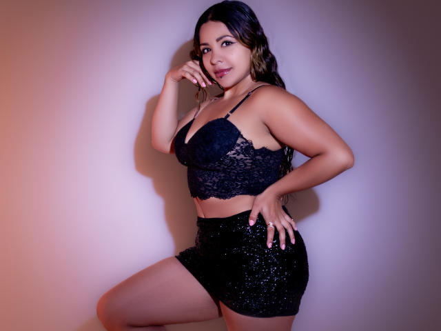 VictoriaLuren with medium tits, wearing a black lace bra and sparkly black mini-skirt, standing with one hand on her hip and the other touching her chin, smiling.