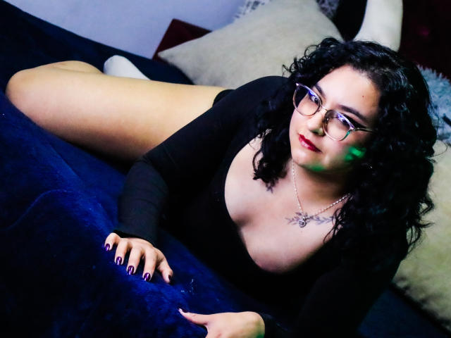 AmberColombo with curly black hair, glasses, black top, and visible tits, lying on blue velvet, legs up, white socks, black nails, necklace, and tattoo, looking at camera.