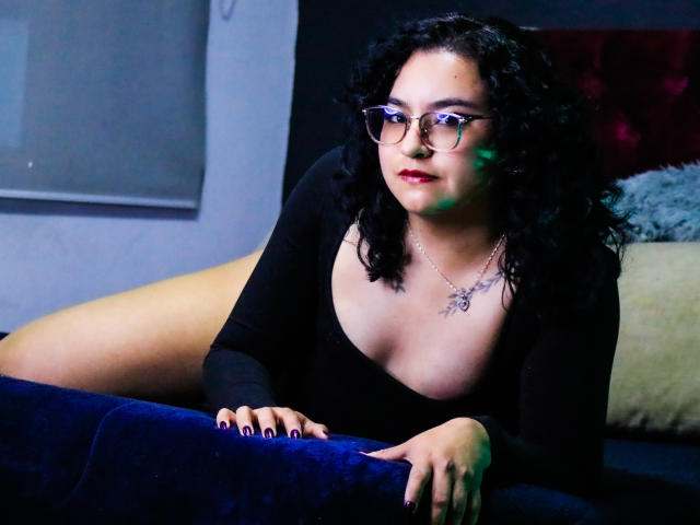 AmberColombo with curly black hair, glasses, and red lipstick, leaning on a blue couch in a black dress, showing her tits, wearing a necklace, with purple nails.