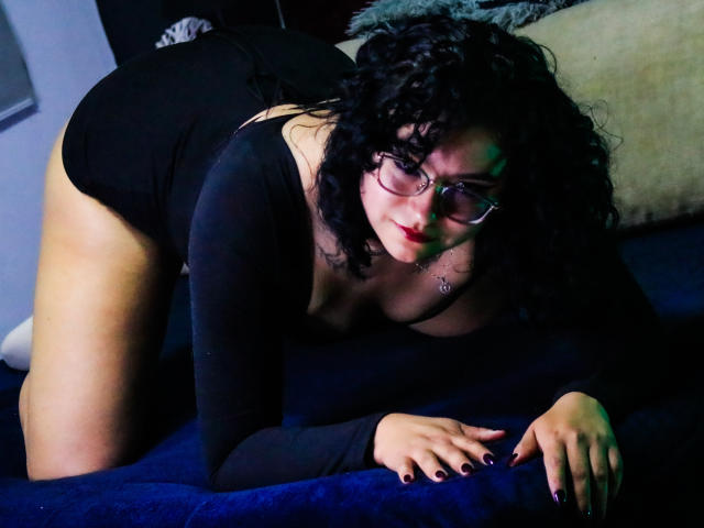 AmberColombo on all fours, wearing black lingerie, glasses, and curly black hair, with large tits and a round ass, on a blue couch, looking down with a slight smile.