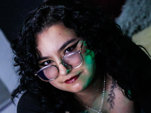 AmberColombo with curly black hair, wearing glasses, smiling with red lipstick, green and blue light on face, tattoo on right chest, necklace, dark top, slightly tilted head.
