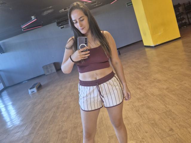 Roussemelinda69 with long black hair, wearing a maroon sports bra and striped white and brown shorts, taking a mirror selfie in a wooden-floored room. Her tits are small.