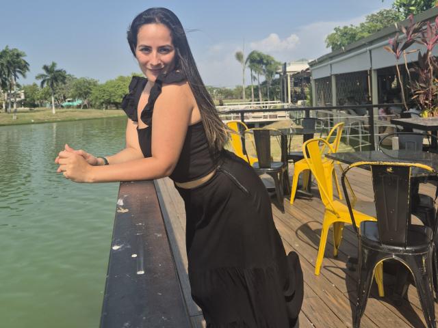 Roussemelinda69 with long dark hair, black ruffled crop top, and black skirt, leaning on wooden railing, smiling, yellow metal chairs, green water, wooden deck, sunny day.