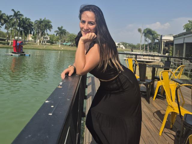 Roussemelinda69 with long dark hair, black sleeveless dress, leaning on wooden railing, hand under chin, yellow chairs and table on right, green water, palm trees, smiling, sunny day.