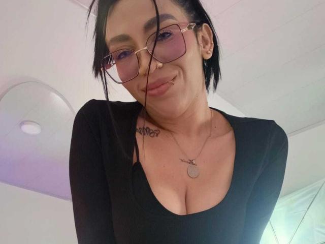 CamilleJuicyHot with black hair, glasses, and nose ring, wearing a black top, showing her tits, smiling, and tattoos on her neck and chest.