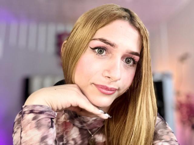 AndyDupont with long blonde hair, pink eyeshadow, and pink lipstick, resting chin on hand, wearing a tie-dye shirt, looking at camera with a slight smile, in selfie.