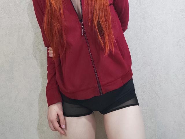 SheritaChieng with long red hair, wearing a red zip-up hoodie and black mesh shorts, exposing her pale thighs and a hint of her pussy. Hands by her sides.