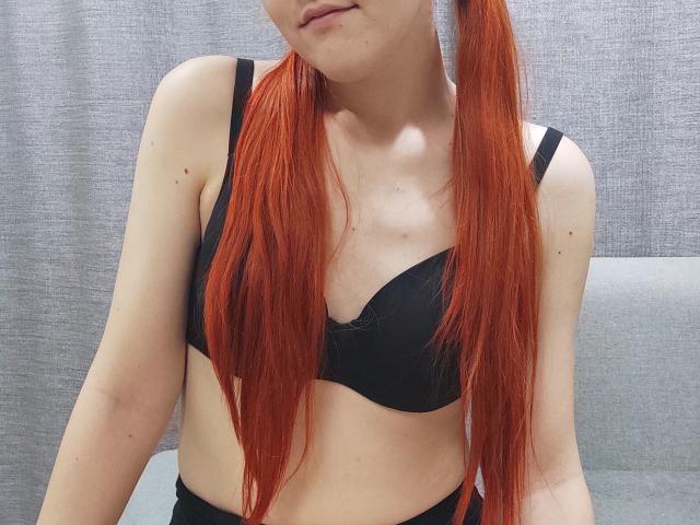SheritaChieng, com longas tranças ruivas, vestindo um sutiã preto, mostrando seus pequenos seios e pele clara, inclinando levemente a cabeça com um sorriso sutil e discreto..