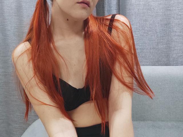 SheritaChieng with red pigtails, wearing a black bra, showing her small tits and pale skin, slightly biting her lower lip, gray curtain in the background.