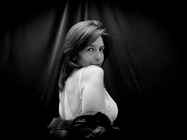 Black-and-white photo of SabinaDeluxe with shoulder-length hair, looking over her shoulder, wearing a white lace bra, and a black leather jacket partially draped. Boobs visible.