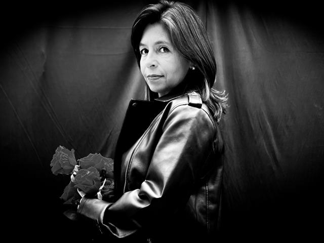 SabinaDeluxe with medium tits, holding roses, wearing a shiny leather jacket, looking to the side, black and white, medium shot, slight smile, wavy hair, dark background.