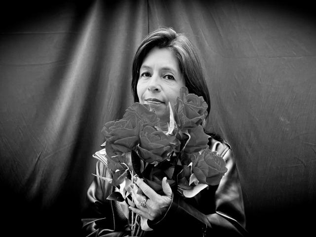 SabinaDeluxe with roses, holding them to her face, wearing a leather jacket, black and white, smiling, ring on her right hand, medium-length dark hair, soft light, gentle expression.