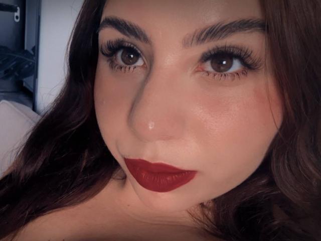 VenusxMusa with dark brown eyes, thick eyelashes, red lipstick, and slightly shiny skin, looking directly at the camera with dark brown, wavy hair framing her face.