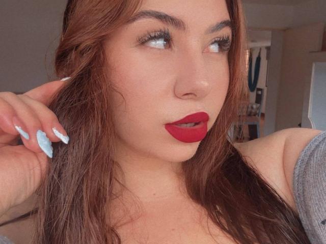 VenusxMusa with red lipstick, long brown hair, light blue nails, looking to the side, gray off-shoulder top, slightly parted lips, soft light, slight blush on her cheeks.