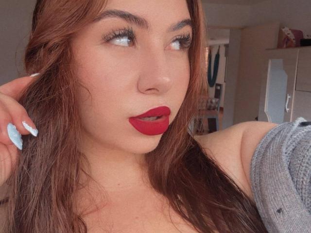 VenusxMusa with red lipstick, long brown hair, blue nails, looking to the side, wearing a grey off-shoulder top, showing her tits.
