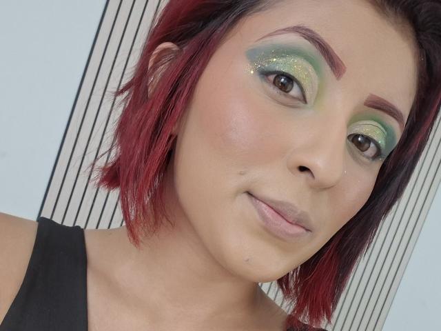 LizBonnet's face with red-dyed hair, green and yellow glittery eyeshadow, brown eyes, and a small beauty mark on her left cheek, wearing a black top.