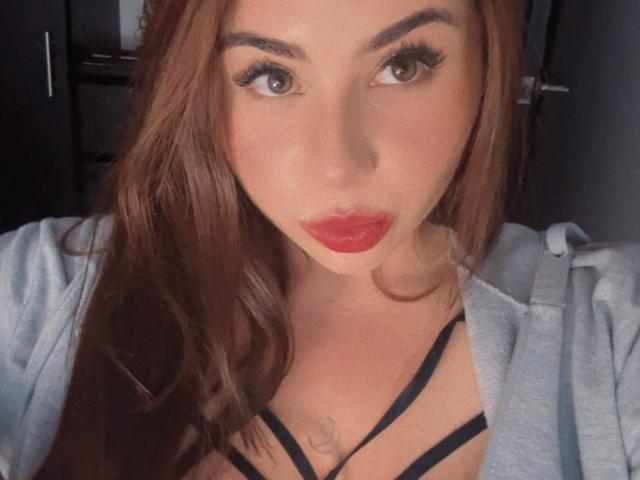 VenusxMusa con rossetto rosso, occhi marroni e lunghi capelli castani, indossa una felpa grigia e un top nero con spalline sottili, mostrando il seno. Il suo trucco è impeccabile..