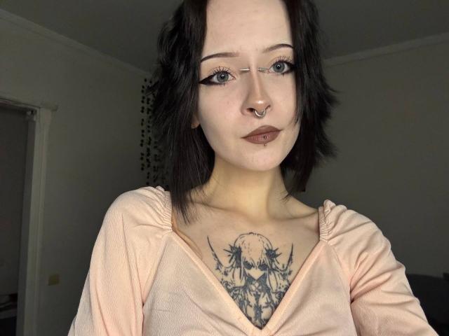 LianeBadenoch with black hair, nose ring, and dark lipstick, wearing a pink top, showing a chest tattoo of a moon and stars, has bold eyeliner and a lip piercing.