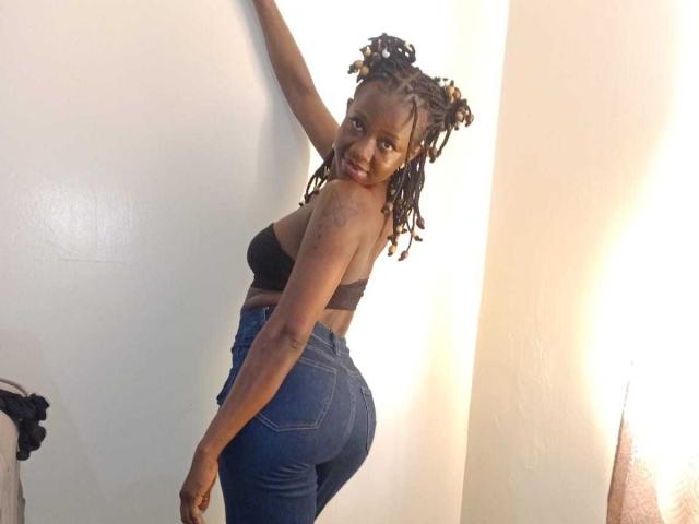 SexyMindset with dark skin and braided hair, wearing a black strap top and blue jeans, looking back with a smile, showing her ass and small tattoo on her arm.