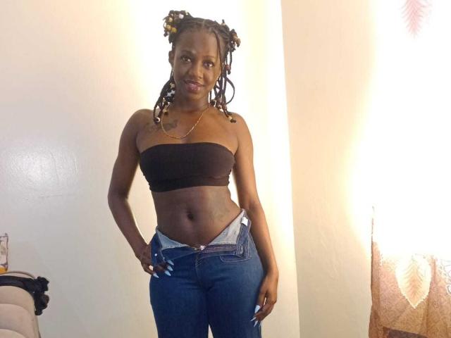 SexyMindset with dark skin, wearing a black strapless top, showing her tits and a flat stomach, blue jeans, and her hand in her pocket, standing with a smile.