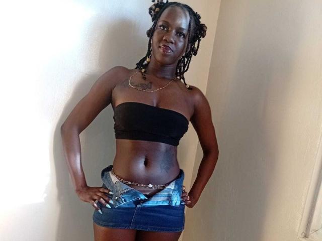SexyMindset has dark skin, small boobs, and wears a black strap top, denim skirt, and unbuttoned jeans with a belt, standing with hands on hips, smiling.