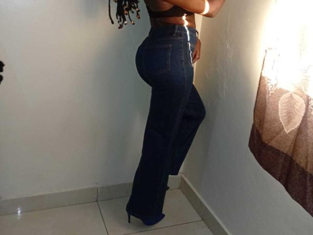 SexyMindset standing sideways, wearing dark blue jeans and blue heels, black bra, showing her ass, with long braided hair, white wall, and brown curtain in the background.