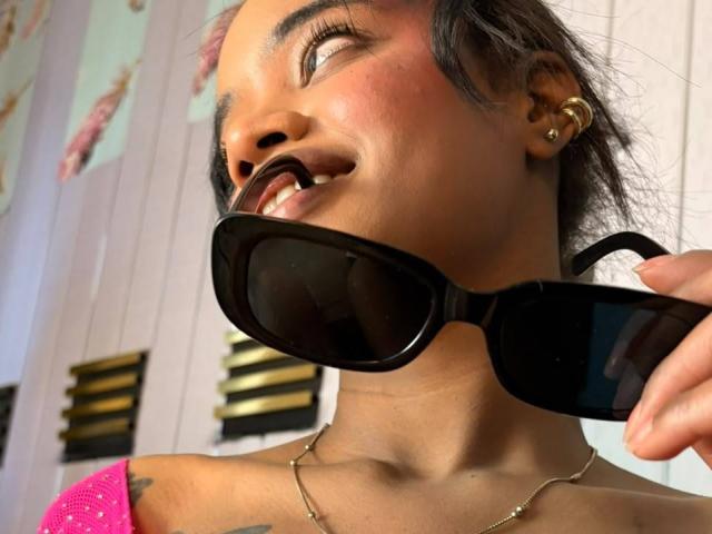 KiaraBrown holding black sunglasses in her mouth, eyes closed, smiling, with pink top and gold earrings, showing tattoo on her shoulder. She's wearing a gold necklace.