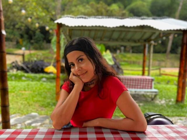 ElectraRoss with dark hair, wearing a red shirt and black headband, resting her chin on her hand, smiling at the camera, sitting at a red and white checkered table.