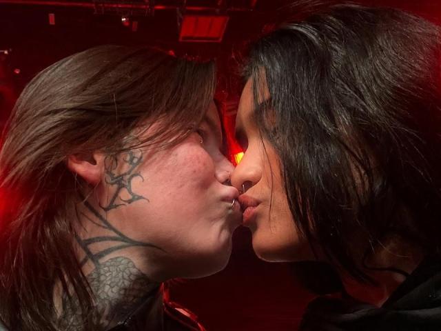 KaliyKatie with tattoos on the side of her face kissing another person with dark, wet hair in a red-lit room, noses touching, lips slightly parted.