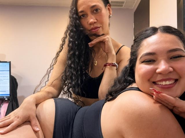CharlotteTiffany with long curly hair, black sports bra, and red bracelet, touching another CharlotteTiffany's ass, who's smiling, wearing black sports bra, and red nails.