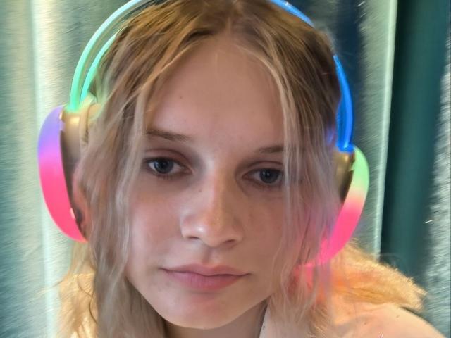 EdyTheViard with neon rainbow headphones, blonde hair, blue eyes, and a neutral expression. She has a fair skin tone and a slightly messy hairstyle.