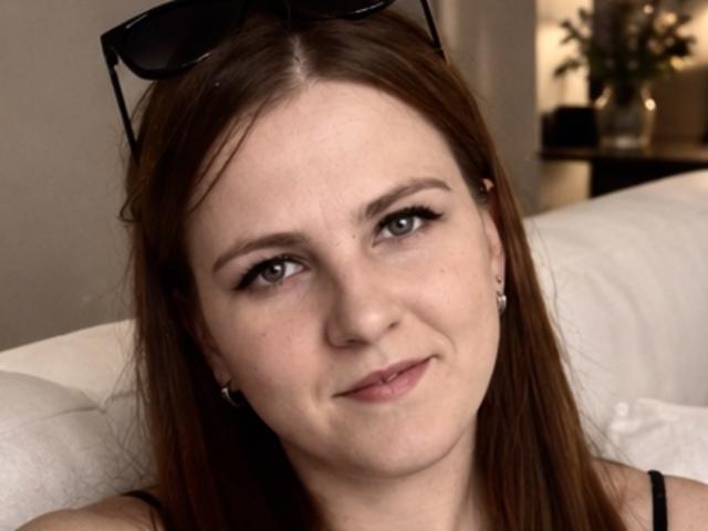 ElizabethGlow has brown hair, light skin, and small tits, wearing black earrings and sunglasses on her head, with a slight smile and subtle makeup.