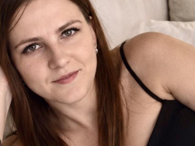 ElizabethGlow has light skin, brown eyes, and long brown hair, wearing a black spaghetti strap top, looking at the camera with a slight smile. She has small earrings.
