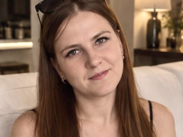 ElizabethGlow with straight brown hair, small tits, wearing black earrings and a sleeveless top, smiling slightly, has glasses on her head, sitting on a white couch.