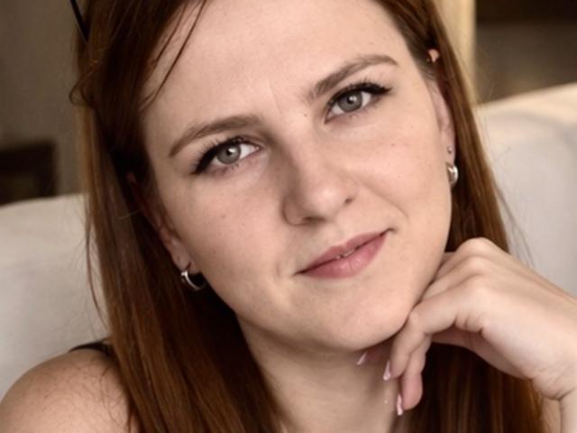 ElizabethGlow has light brown hair, green eyes, and a slight smile. She's wearing small earrings, with her hand under her chin, and has a cute, relaxed look.