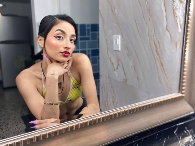 LunaixMoon, avec ses cheveux foncés, son rouge à lèvres rouge, son soutien-gorge floral jaune, le menton appuyé sur sa main, regardant dans le miroir, montrant de petites seins, un tatouage au poignet, des ongles violets, un reflet sur un mur en marbre..