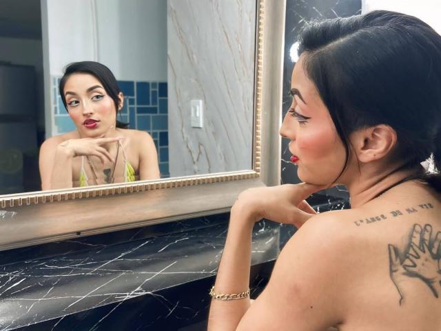 LunaixMoon with dark hair, red lipstick, and tattoos, looks in a mirror, resting her chin on her hand, showing her bare shoulders and black marble countertop.