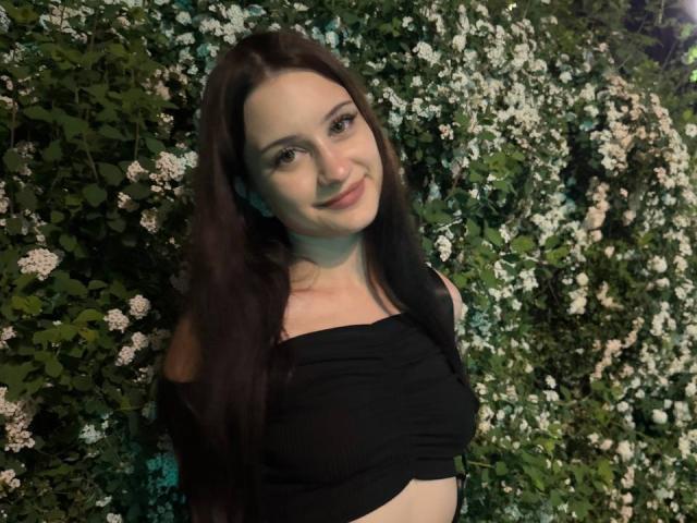 EleanorEvanchyk with long brown hair, smiling, wearing a black off-shoulder top, exposing her small tits, standing in front of a green, white-flowered wall.