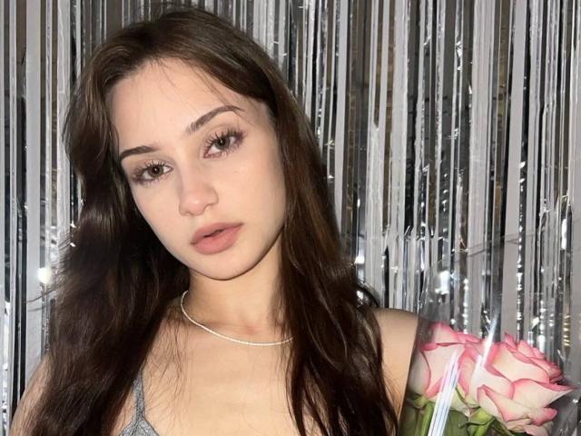 EleanorEvanchyk with long brown hair, wearing a thin silver necklace, and holding pink roses, has soft pink lips and subtle makeup, looking slightly bored.
