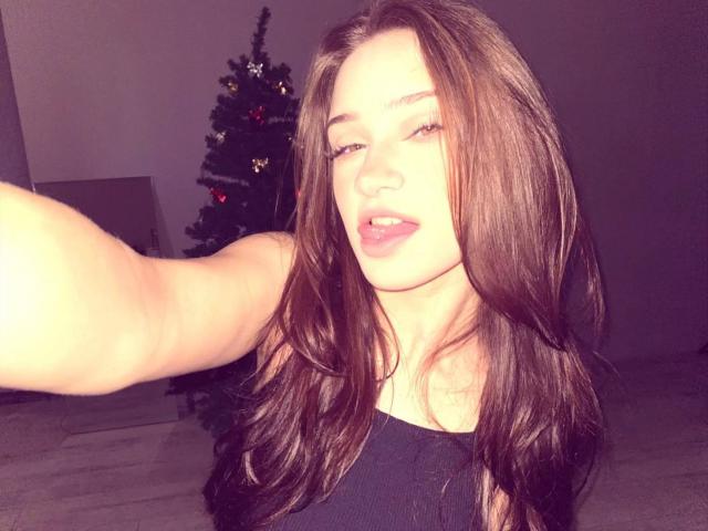 EleanorEvanchyk with long brown hair, wearing a black top, taking a selfie, showing her slightly open mouth and raised arm, with a Christmas tree in the background.