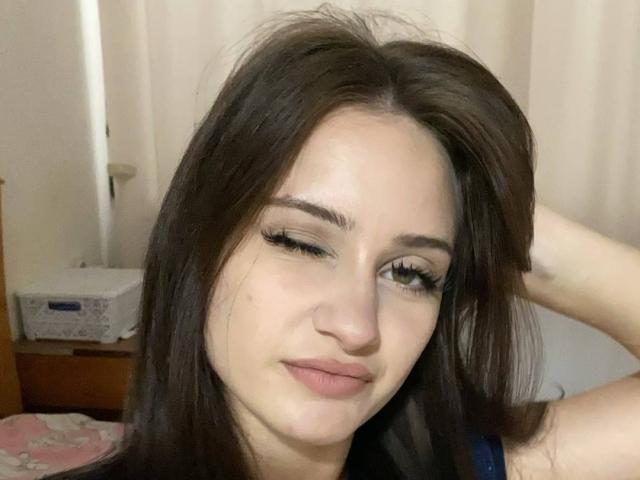 EleanorEvanchyk with long brown hair, winking, has small tits, fair skin, and long eyelashes, sitting with hand in hair, smiling slightly, in a bedroom.