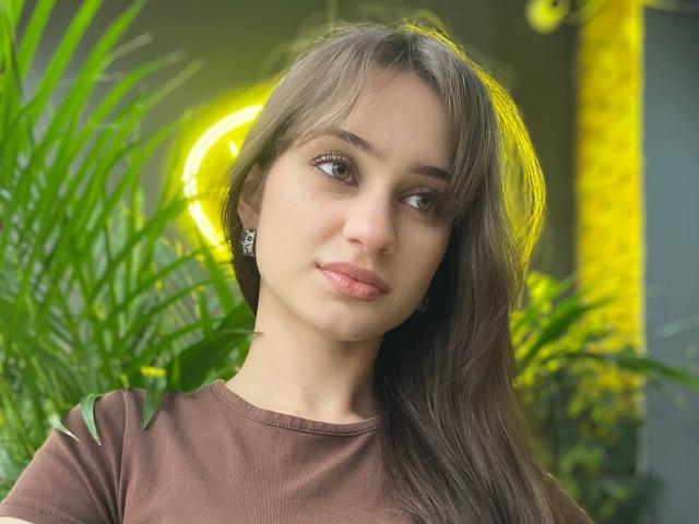 EleanorEvanchyk has brown hair, green eyes, and a subtle smile. She's wearing a brown shirt with green leaf accents. Her tits are small, and she's looking to the side.