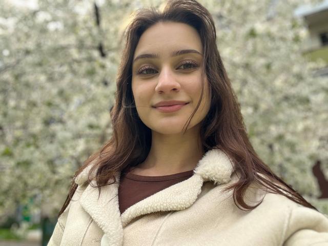 EleanorEvanchyk with brown hair, green eyes, and light skin, wearing a beige fleece jacket over a brown shirt, smiling softly. Boobs not visible, ass not visible, pussy not visible.