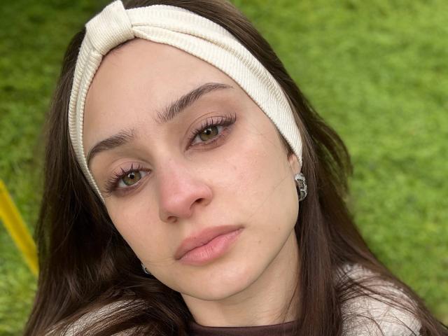EleanorEvanchyk with brown eyes, long brown hair, white headband, and pink lips, showing off her small nose and slightly upturned ass-adjacent lip curve. She's wearing a dark shirt.