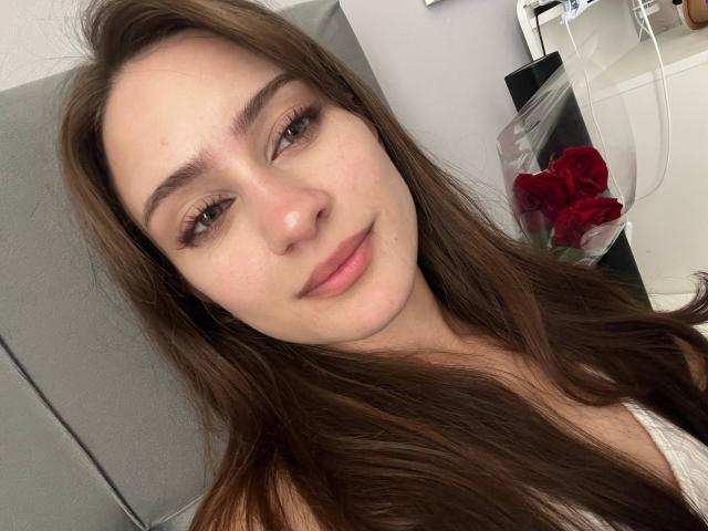 EleanorEvanchyk with long brown hair, brown eyes, and a slight smile, wearing a white top, has red roses in a clear bag on a bathroom counter.
